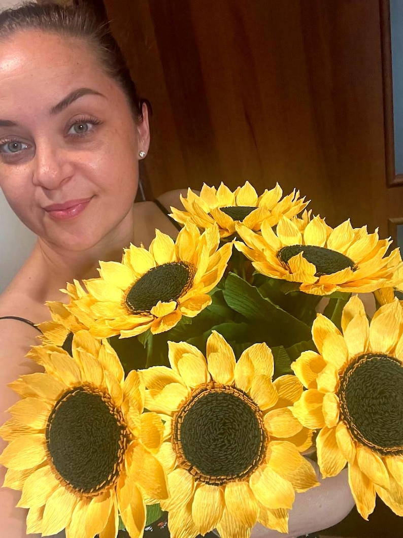 Peut inclure: Un bouquet de grands tournesols en papier avec des p&eacute;tales jaune vif et des centres brun fonc&eacute;. Les fleurs ont des feuilles de papier vertes. Le bouquet est tenu par une personne.