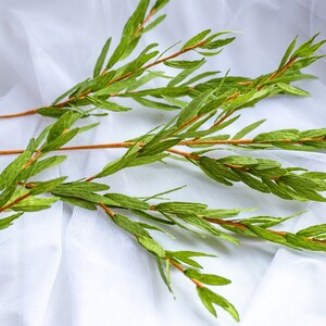 Op de afbeelding: Een bos groene papieren bladeren op een witte stoffen achtergrond. De bladeren zijn in een natuurlijk, vloeiend patroon gerangschikt, wat een gevoel van organische schoonheid cre&euml;ert.
