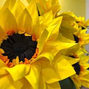 Puede incluir: Un grupo de grandes girasoles hechos a mano con p&eacute;talos de color amarillo vibrante y un centro marr&oacute;n oscuro texturizado. Los p&eacute;talos est&aacute;n dispuestos en capas y son puntiagudos, creando una apariencia completa y tridimensional.