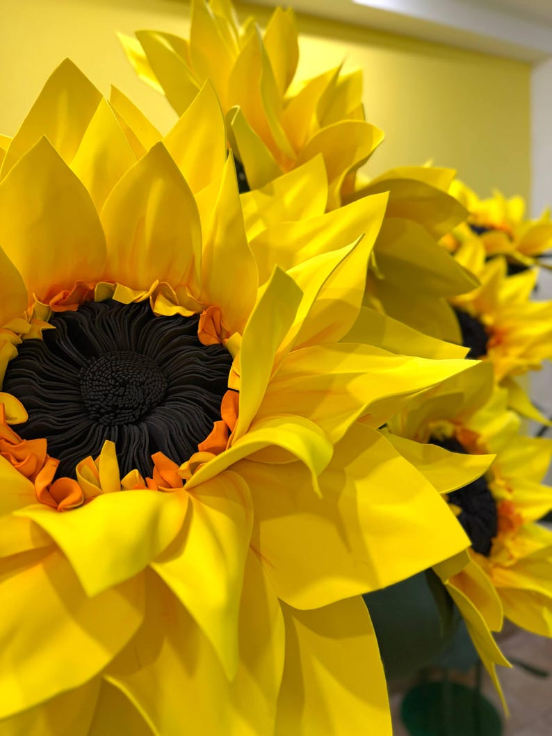 Puede incluir: Un grupo de grandes girasoles hechos a mano con p&eacute;talos de color amarillo vibrante y un centro marr&oacute;n oscuro texturizado. Los p&eacute;talos est&aacute;n dispuestos en capas y son puntiagudos, creando una apariencia completa y tridimensional.