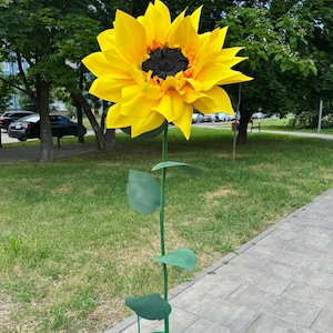Puede incluir: Una gran escultura decorativa de girasol de gran tama&ntilde;o con p&eacute;talos de color amarillo brillante y un centro negro texturizado. La flor est&aacute; unida a un tallo de metal verde delgado con tres hojas verdes, apoyada sobre una base circular de metal verde.