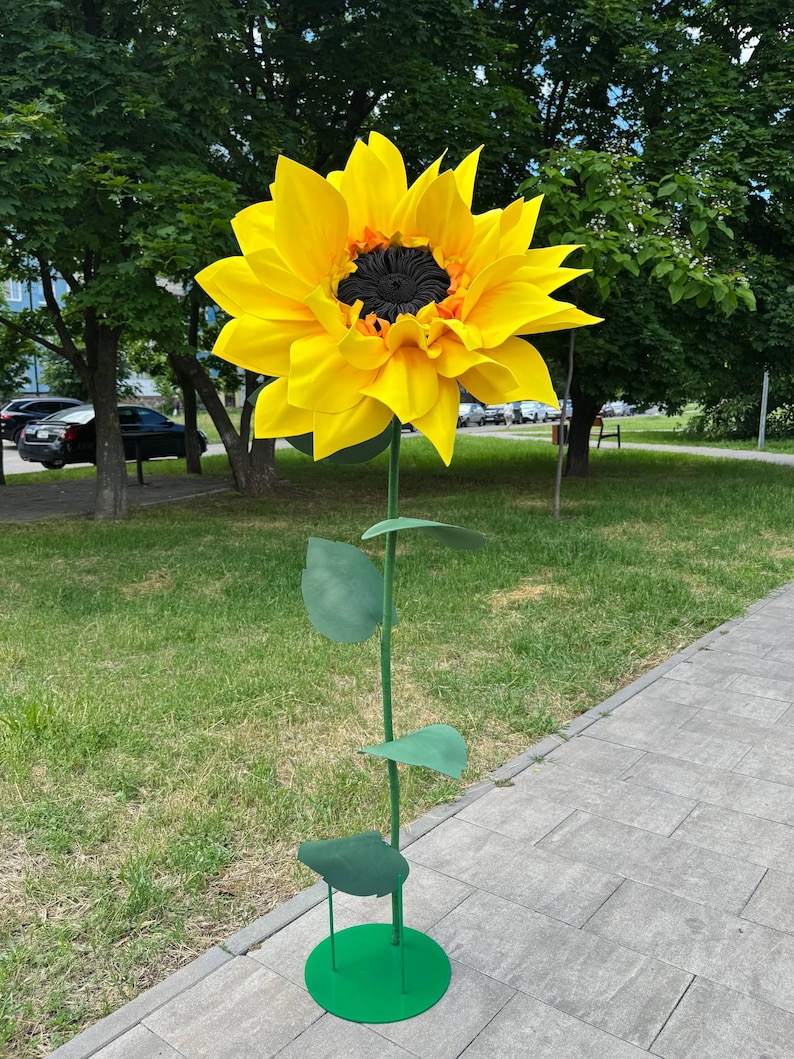 Puede incluir: Una gran escultura decorativa de girasol de gran tama&ntilde;o con p&eacute;talos de color amarillo brillante y un centro negro texturizado. La flor est&aacute; unida a un tallo de metal verde delgado con tres hojas verdes, apoyada sobre una base circular de metal verde.