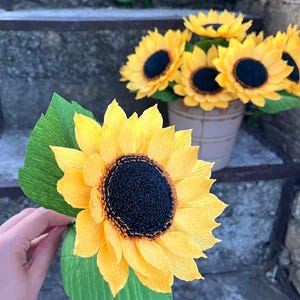 Peut inclure: Gros plan sur un tournesol en papier fait &agrave; la main avec des p&eacute;tales jaunes et un centre sombre, tenu dans une main. D'autres tournesols sont dans un petit pot beige. Des feuilles de papier vertes sont attach&eacute;es &agrave; la fleur.