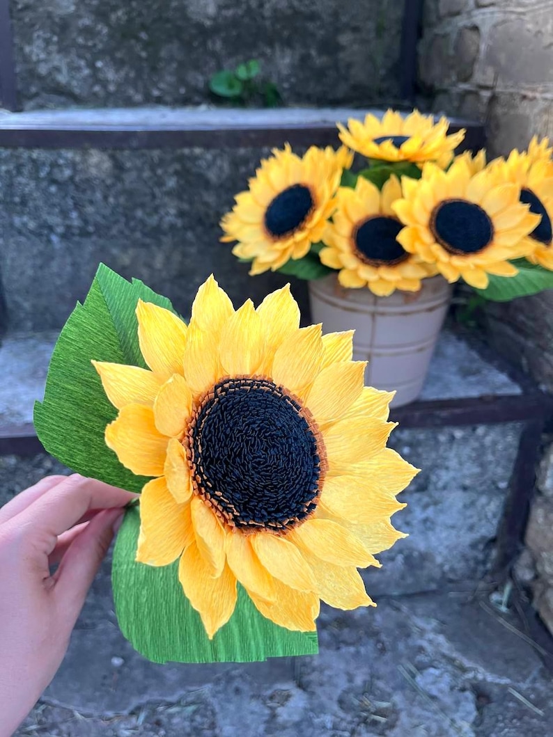 Peut inclure: Gros plan sur un tournesol en papier fait &agrave; la main avec des p&eacute;tales jaunes et un centre sombre, tenu dans une main. D'autres tournesols sont dans un petit pot beige. Des feuilles de papier vertes sont attach&eacute;es &agrave; la fleur.