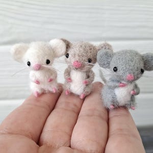 May include: Three miniature felt mice in white, tan, and gray, each with pink paws and noses, black eyes, and delicate whiskers. The mice are standing upright, showcasing their small size and detailed craftsmanship.