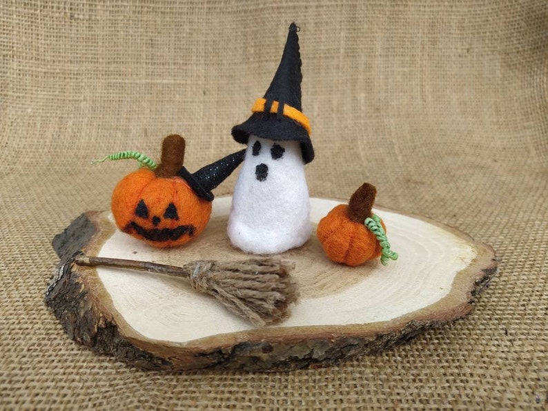 May include: A felt ghost wearing a black witch hat with an orange band, holding a small broom. Two orange felt pumpkins are on a wooden slice.