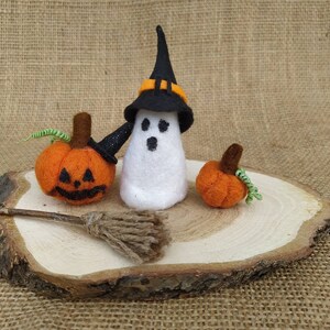 May include: A white felt ghost wearing a black witch hat with an orange band, holding a small black broom. Two orange felt pumpkins with green stems are on a wooden slice.