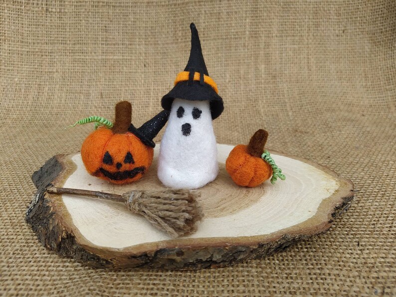 May include: A white felt ghost wearing a black witch hat with an orange band, holding a small black broom. Two orange felt pumpkins with green stems are on a wooden slice.