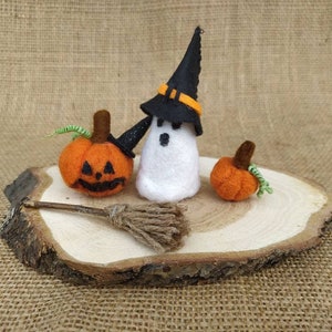 May include: A white felt ghost wearing a black witch hat with an orange band, holding a small brown broom. Two orange felt pumpkins with green stems are on a wooden slice.