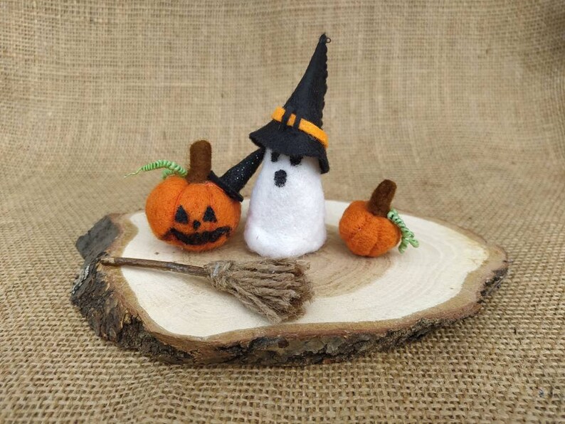 May include: A white felt ghost wearing a black witch hat with an orange band, holding a small brown broom. Two orange felt pumpkins with green stems are on a wooden slice.