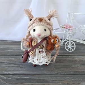 以下が含まれることがあります： 茶色のニットのフクロウの帽子と茶色のニットのカーディガンを着た、小さな白いフェルトのマウス。マウスは小さな茶色のフェルトのフクロウと茶色の革のバッグを持っています。マウスは灰色の木製の表面に立っています。