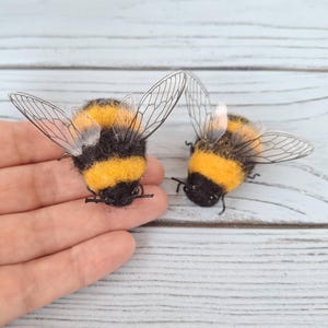 Pode incluir: Dois abelhões de lã feltrada com corpos felpudos de riscas pretas e amarelas. Cada abelha apresenta asas delicadas, translúcidas e nervuradas feitas de um material fino, além de pequenas antenas pretas e um aspeto texturizado.