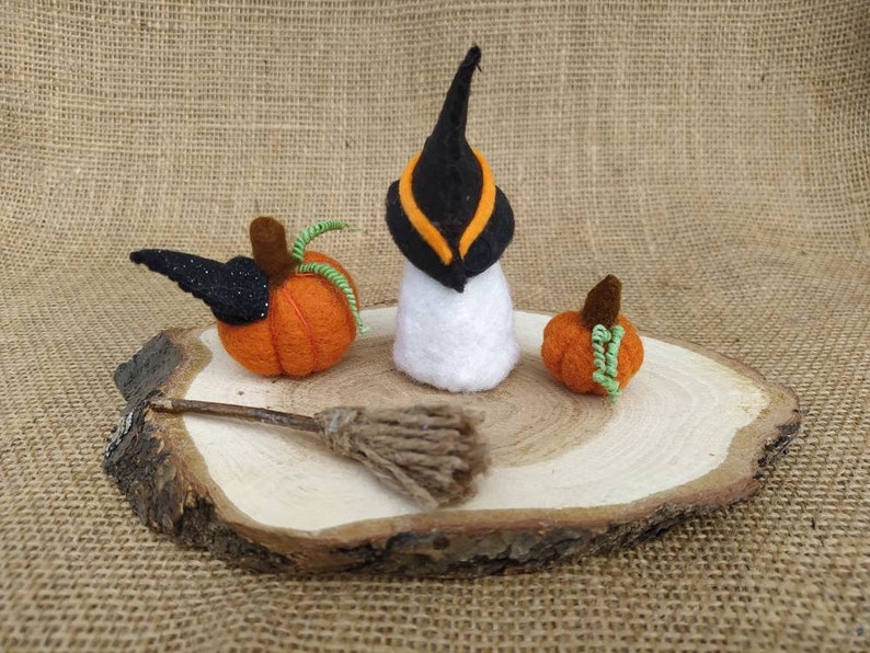 May include: A felt witch with a black hat and orange trim, a broom, and two orange pumpkins with green stems on a wooden slice.