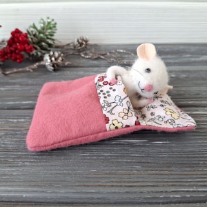 Puede incluir: Un juguete de ratón de fieltro blanco con nariz y orejas rosas, acurrucado en una cama de fieltro rosa con una almohada estampada floral. El ratón tiene ojos negros y una nariz rosa. La cama es de color rosa claro.