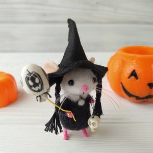May include: A white felt mouse wearing a black witch hat and holding a black and white jack-o-lantern balloon. The mouse is wearing a black dress and has a skull charm on its wrist.
