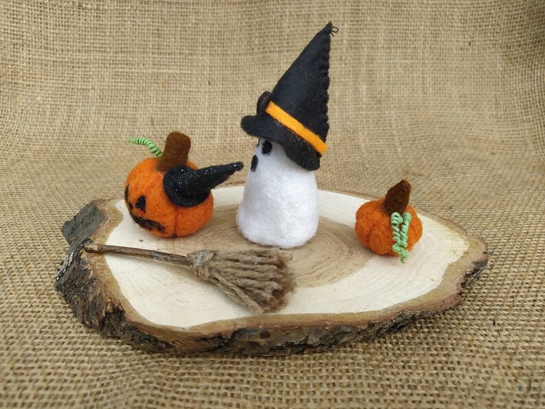 May include: Felt Halloween decorations on a wooden slice. A white ghost wearing a black witch hat with an orange stripe, a small orange pumpkin, and a larger orange pumpkin with a black witch hat.