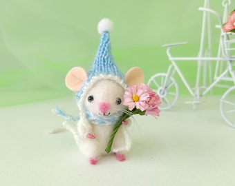 May include: A small white mouse wearing a blue knitted hat and scarf holds a bouquet of pink flowers. The mouse is sitting on a white surface with a green background.