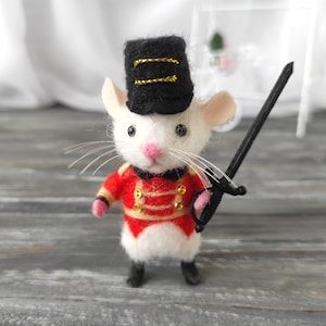 Pode incluir: Um rato de feltro branco usando um chapéu preto e dourado e um uniforme vermelho e dourado, segurando uma espada preta. O rato está em pé sobre uma superfície de madeira.