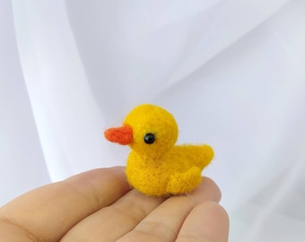 May include: A small yellow felt duck with an orange beak and black eyes. The duck is sitting on a hand.