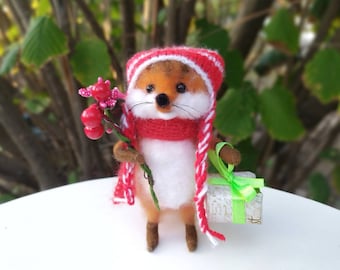 Puede incluir: Un pequeño zorro blanco y naranja que lleva un gorro y una bufanda a rayas rojas y blancas, sosteniendo una rama de bayas rojas y un regalo envuelto con un lazo verde.