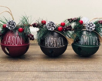 May include: Three Christmas bell ornaments in burgundy, black, and green, each with a name in white script: "Jason", "Hershel", and "The Smith's". The bells are decorated with pine, berries, and pinecones, and have twine hangers.