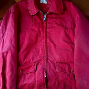 May include: A red zip-up jacket with two large pockets and a smaller pocket on the left chest. The jacket has a label that reads "The Original " and "The " in a smaller font.