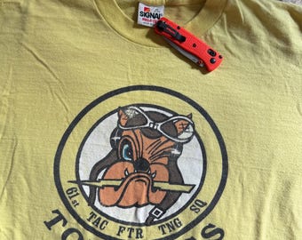 Puede incluir: Camiseta amarilla con un dibujo animado de un bulldog con gafas de aviador y un gráfico "61st TAC FTR TNG SQ TOP DOGS".