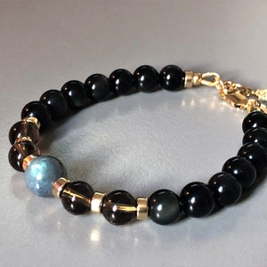 May include: A beaded bracelet featuring black, brown, and blue stones. The bracelet has a gold clasp and a small gold charm.