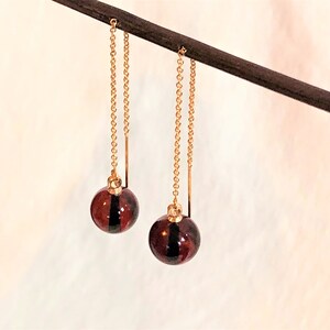 Puede incluir: Un par de pendientes de aro dorados con esferas de vidrio rojo oscuro. Las esferas tienen una franja negra en el centro.