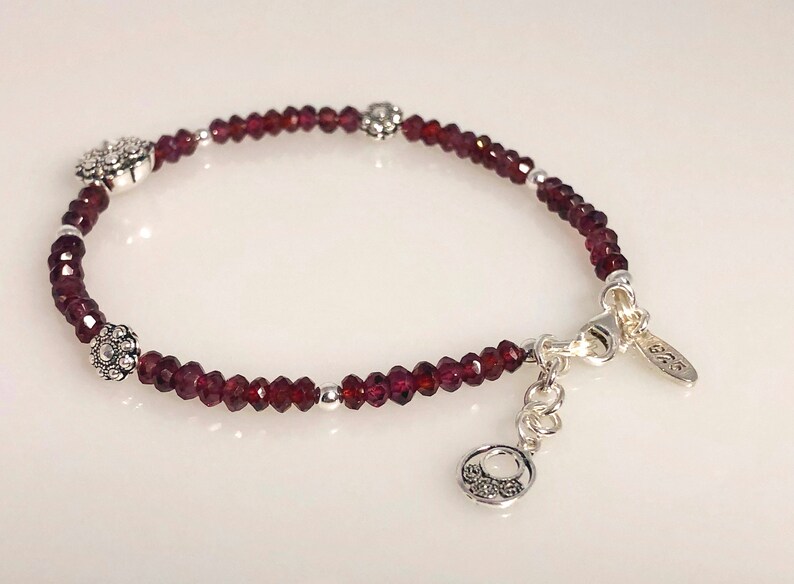 May include: A silver bracelet with a clasp and three silver flower charms. The bracelet is made with small, dark red garnet beads.