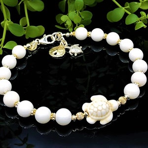 May include: White shell bead bracelet with a gold turtle charm and a gold seashell charm. The bracelet is on a black background.
