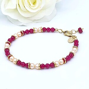 May include: A delicate gold-toned bracelet featuring alternating sections of small, round, deep red beads and creamy white pearls. The bracelet is accented with gold-colored spacers and a small, round charm near the clasp. The background is white with a white flower.