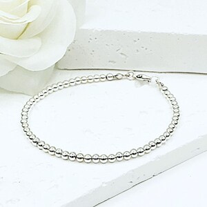 Puede incluir: Una delicada pulsera de cuentas plateadas con un cierre seguro. La pulsera está compuesta por pequeñas cuentas redondas y espaciadas uniformemente. La joya se exhibe sobre una superficie blanca, con una flor blanca en el fondo.