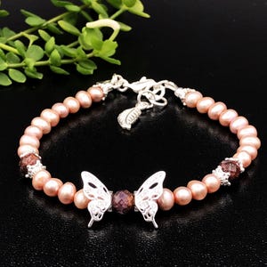 May include: A delicate bracelet featuring a silver butterfly charm with a pink pearl and garnet bead accents. The bracelet is made with small pink pearls and a silver clasp.