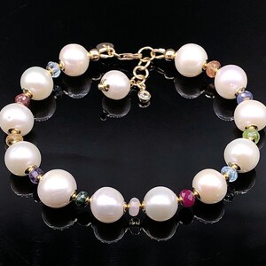 May include: A pearl bracelet with large, round, white pearls interspersed with smaller, colorful gemstones. The bracelet has a gold clasp and gold accents between the pearls and gemstones. The background is black.