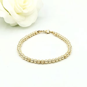 Puede incluir: Pulsera dorada de cuentas con cierre, exhibida sobre una superficie blanca. La pulsera está compuesta por pequeñas cuentas redondas. Una rosa blanca se encuentra en el fondo.
