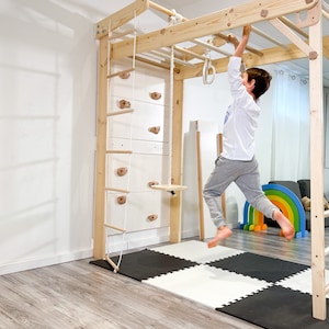 May include: A wooden indoor climbing frame with a ladder, climbing wall, and monkey bars. A child is hanging from the monkey bars. The structure is made of light-coloured wood and is set up on a black and white floor.