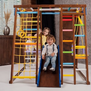 May include: A wooden indoor playset with a climbing wall, a slide, a ladder, and a swing. Three children are playing on the playset. The playset is painted in bright colors, including yellow, blue, green, and red.