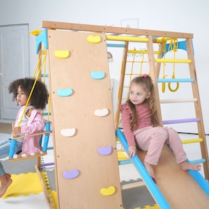 May include: A wooden indoor playground with a climbing wall, slide, swing, and ladder. The playground features colorful accents and is designed for children. Two children are playing on the playground.