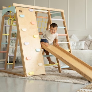 May include: A wooden indoor playset with a climbing wall, ladder, and slide. A child is playing on the slide. The playset is made of natural wood and has a light wood finish.
