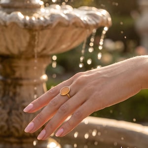 May include: A gold ring featuring a smooth, oval-shaped, translucent orange stone set in a simple metal band. The ring is worn on a finger, showcasing the polished finish of the gemstone and the metallic luster of the band in natural light.
