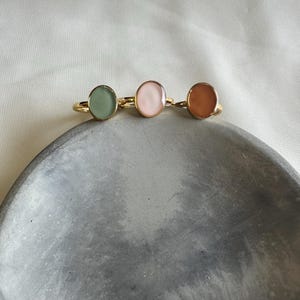 May include: Three gold-toned rings featuring smooth, oval-shaped cabochons in soft pastel colors. The left ring has a sage green stone, the center ring has a pale pink stone, and the right ring has a warm terracotta orange stone, all set in thin metal bands.