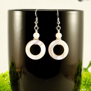 May include: A pair of white shell earrings with a silver hook and a white bead. The earrings are hanging from a black mug.