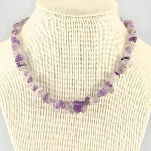 May include: A necklace made of small, irregular amethyst stones. The purple and white stones are strung together, creating a natural, organic look. The necklace is displayed on a beige jewelry bust.