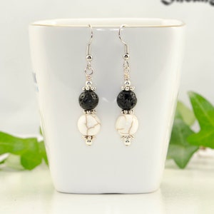 May include: A pair of silver dangle earrings with black lava rock and white howlite beads. The earrings are hanging from a white ceramic mug.