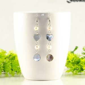 May include: Long drop earrings with a silver chain. Each earring has a heart-shaped, iridescent grey charm, a round grey charm, and small white pearls. The earrings are shown against a white background.