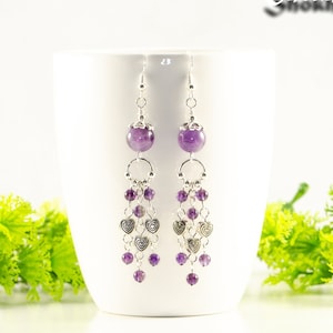 May include: A pair of silver dangle earrings with amethyst gemstones and silver heart charms. The earrings are hanging from a white mug.