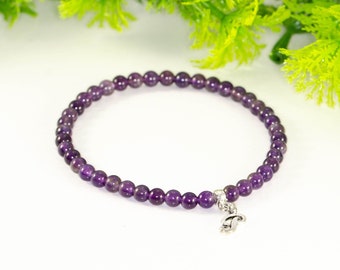 Könnte beinhalten: Ein Armband aus kleinen, runden, tiefvioletten Amethystperlen. Es hat einen silberfarbenen Anhänger. Der Hintergrund ist weiß mit grünem Laub.