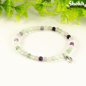 May include: A delicate bracelet made with small, round beads in shades of green, white, and purple. The bracelet has a silver charm in the shape of a circle.