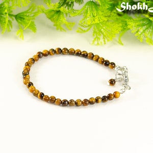 May include: A beaded bracelet featuring small, round tiger's eye stones in shades of brown and gold. The bracelet has a silver-colored clasp and is displayed on a white surface with green foliage in the background. The jewelry is delicate and elegant.